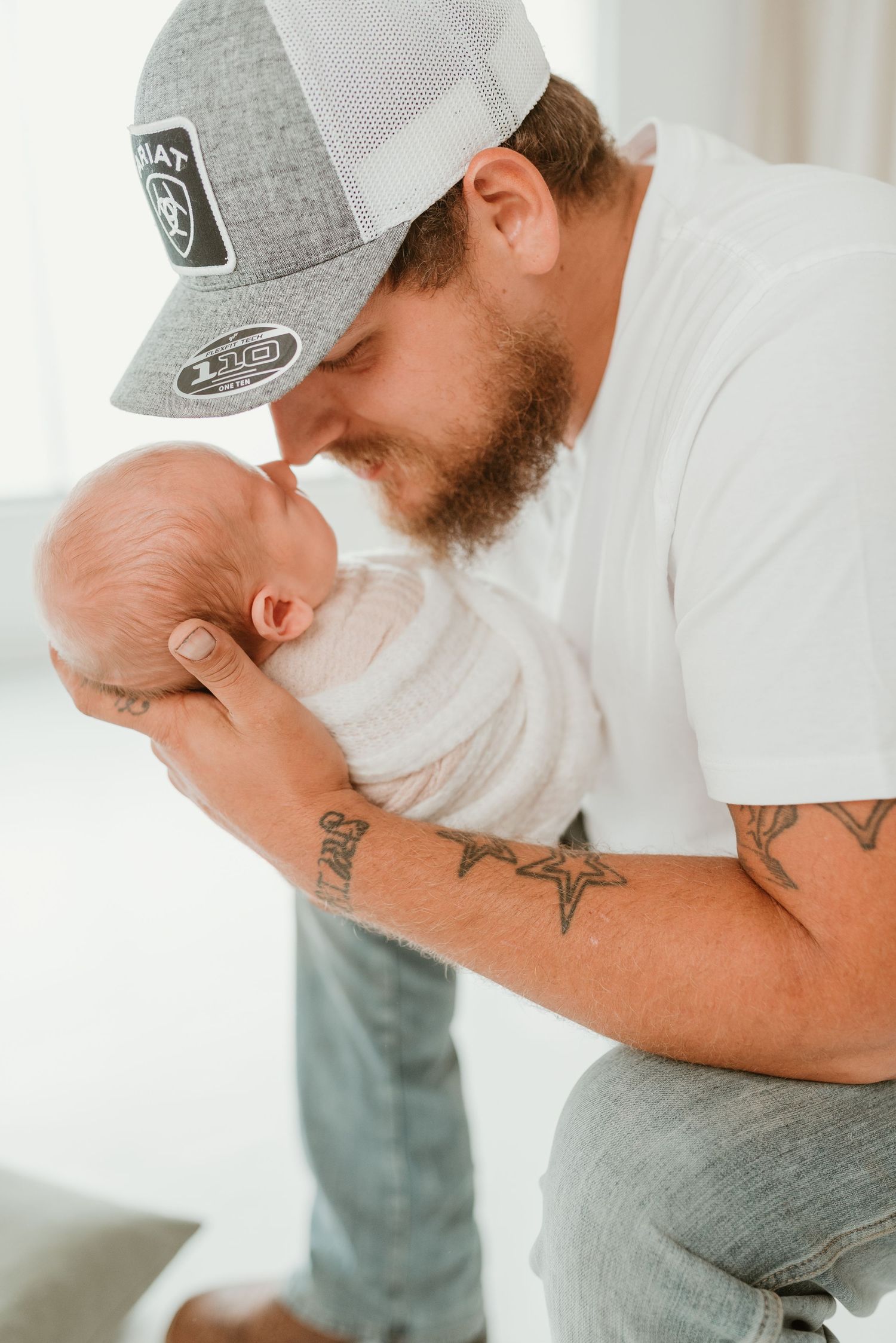 Tattooed father in gray cap tenderly touching foreheads with his swaddled newborn baby.
