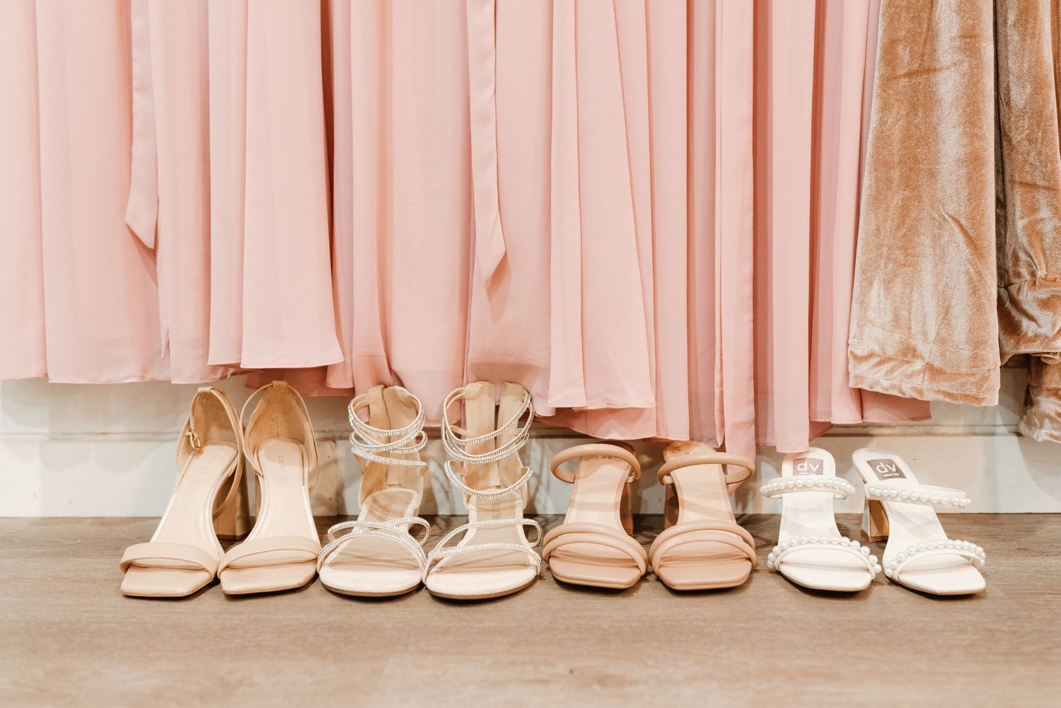 Pink bridesmaid dresses hanging with nude strappy heels arranged neatly on floor in bridal boutique display.