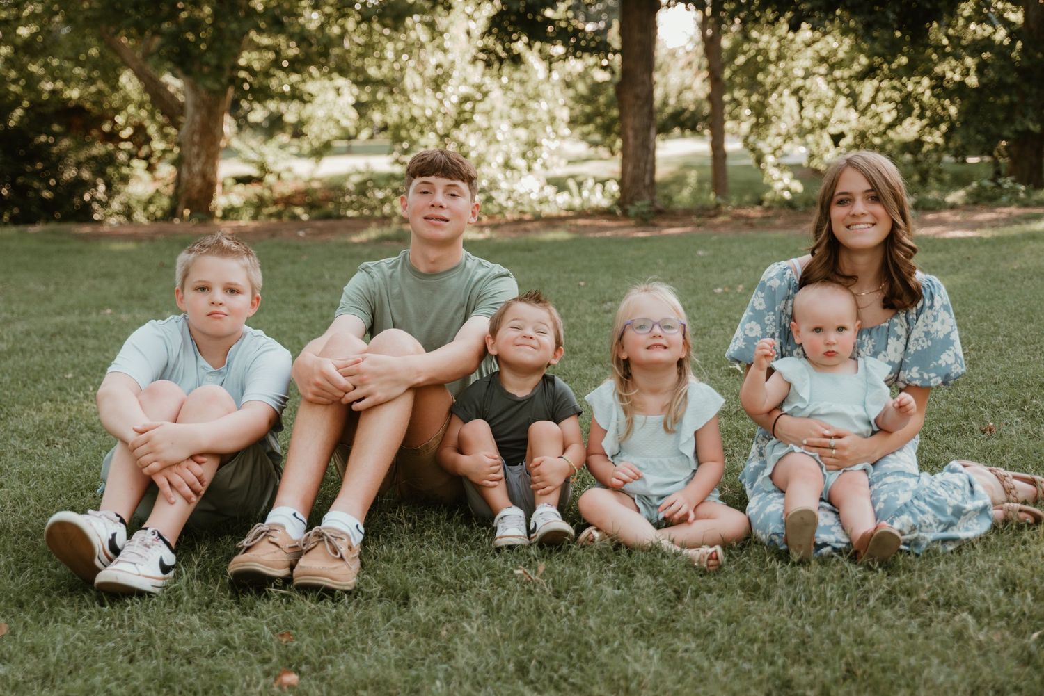 Five children and a young woman sitting together on grass for a family portrait outdoors.
