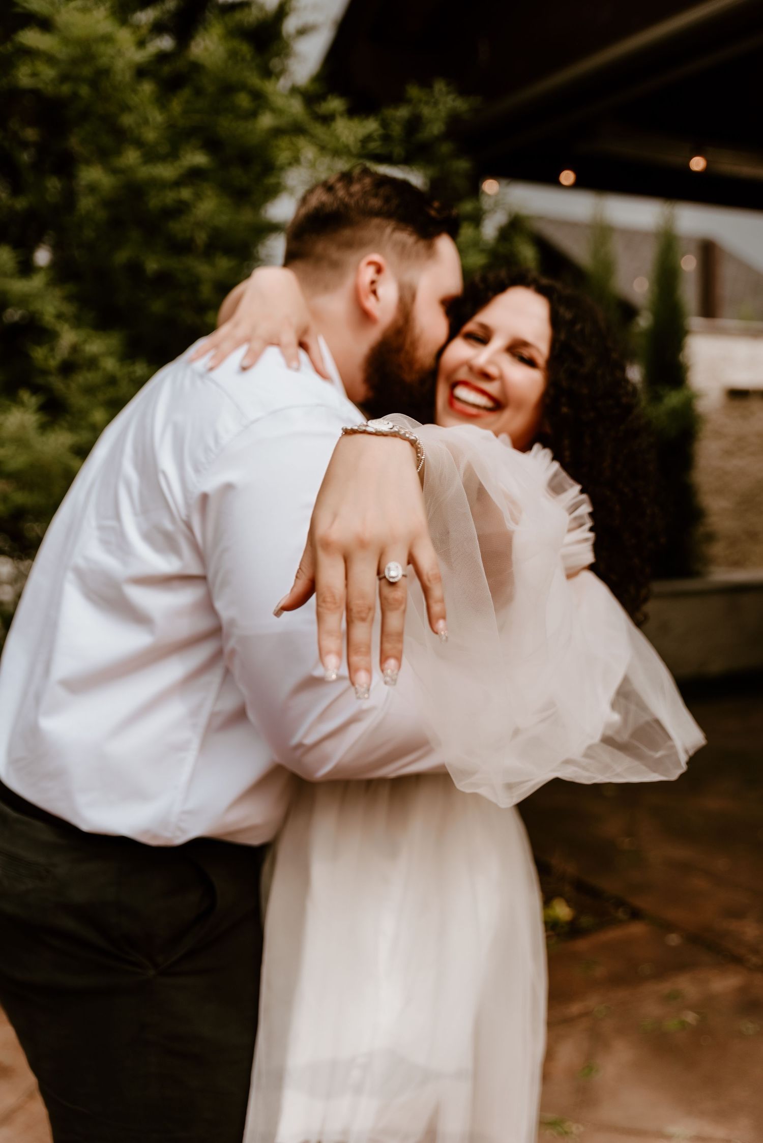 Joyful bride showing off her engagement ring while being embraced by her groom outdoors.