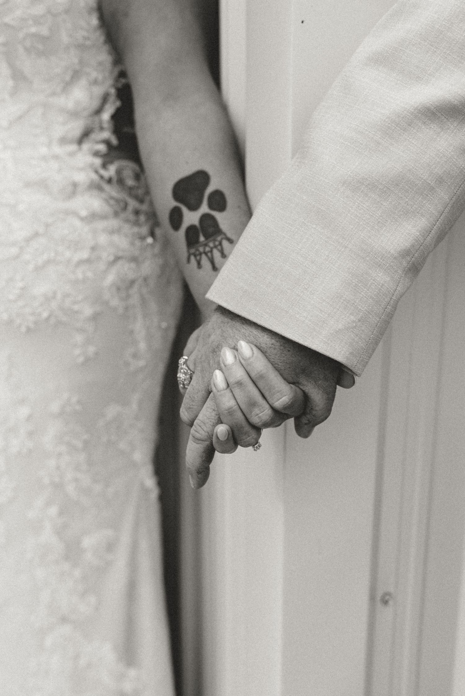 Black and white close-up of bride and groom holding hands revealing paw print tattoo and diamond engagement ring.