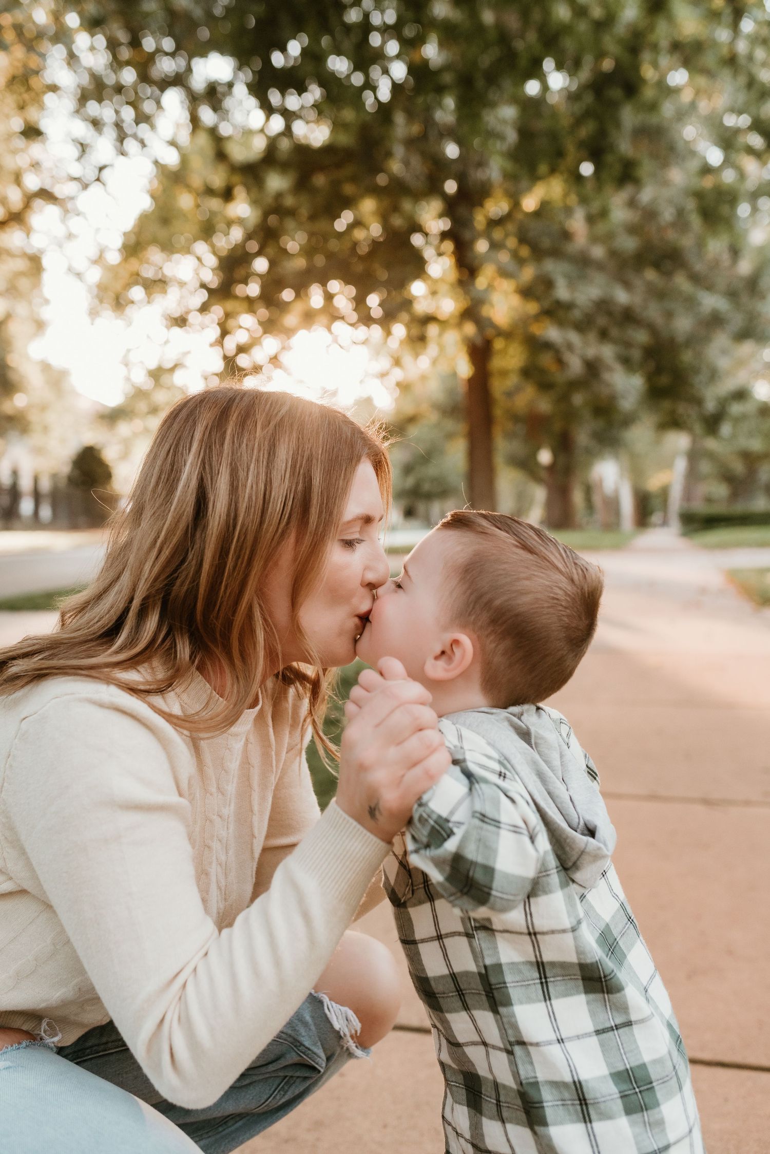 A young mother shares a sweet kiss with her toddler son outdoors in warm golden sunlight.