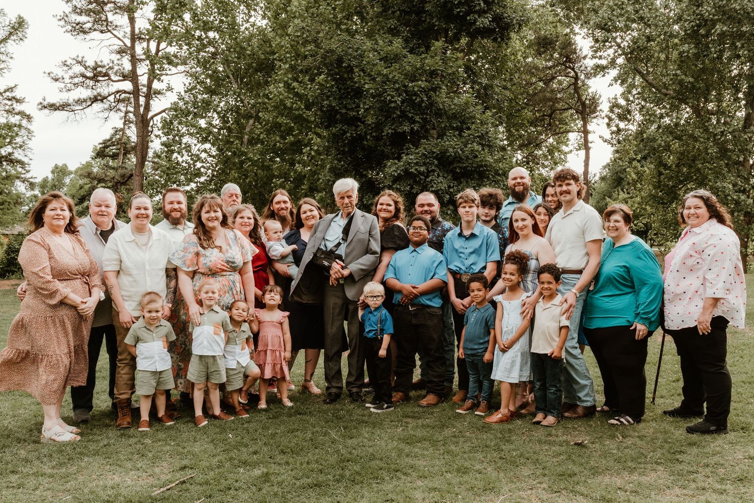 Large multigenerational family group portrait gathered outdoors on grass with trees behind them.