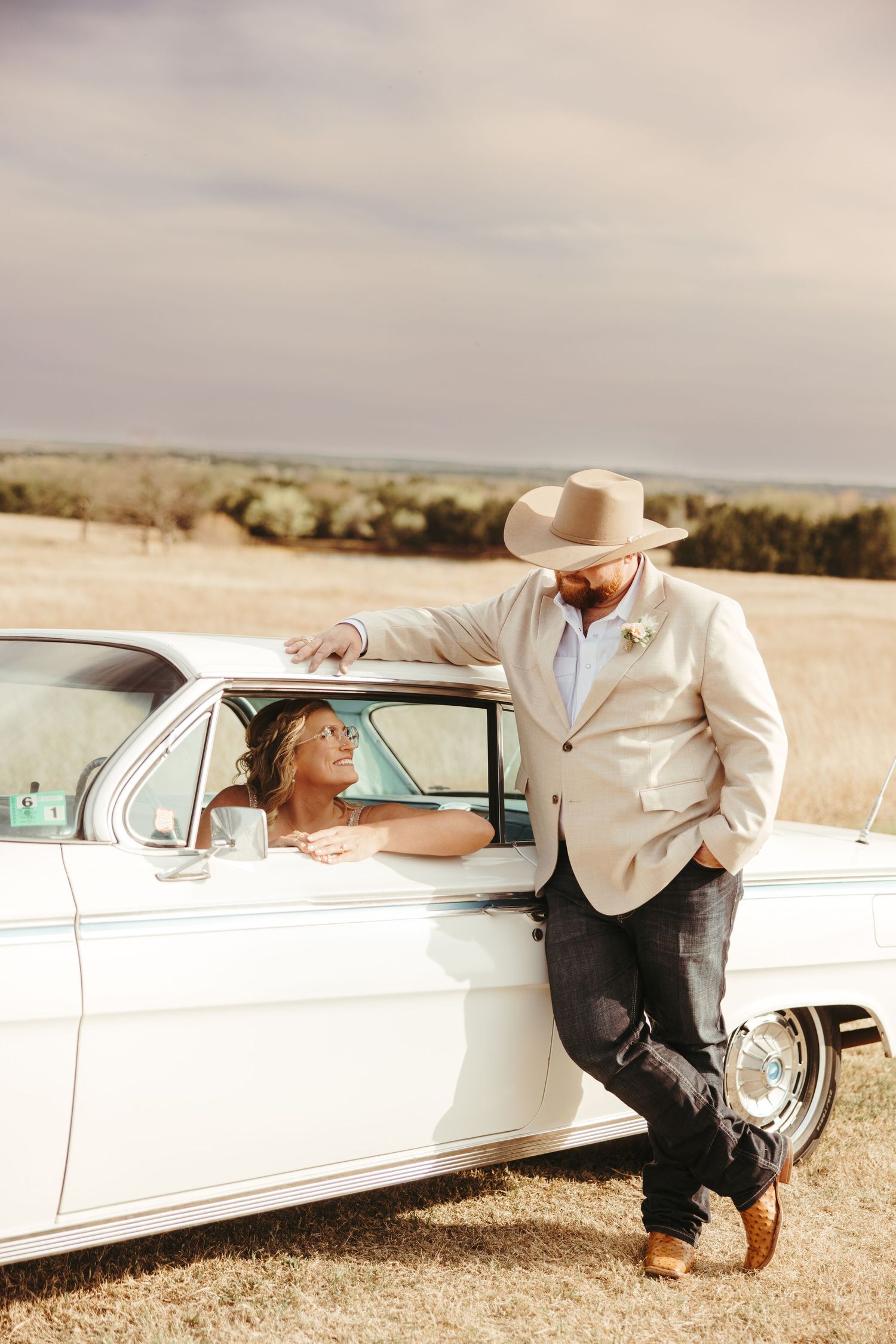 Groom in cowboy hat leans on vintage white car while bride smiles from inside the window.