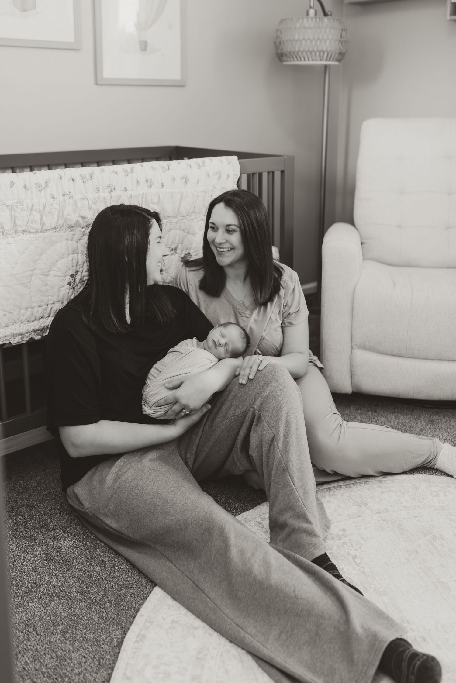 Two women sit on a nursery floor smiling at each other while one holds a newborn baby, black and white.