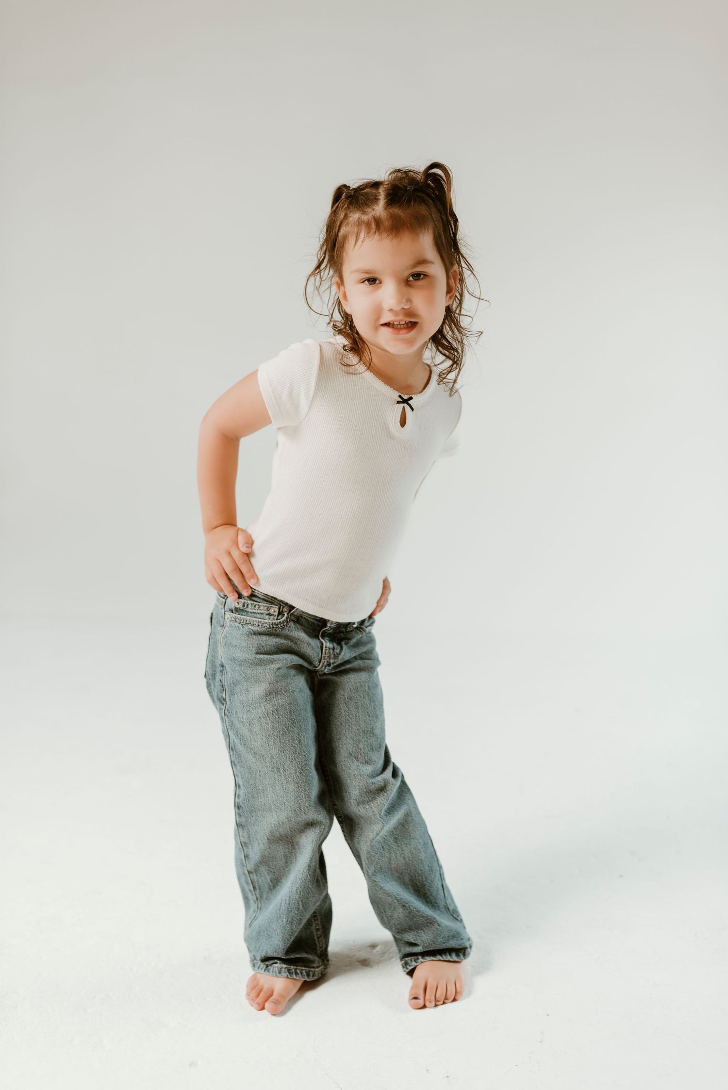 A confident young girl in white tee and oversized jeans poses barefoot on a white background.