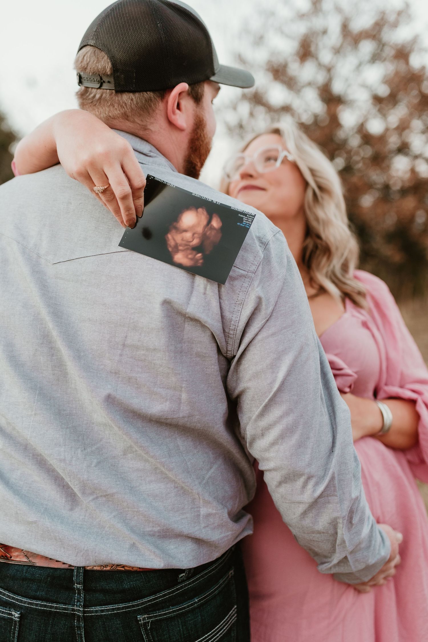 Couple embracing outdoors, woman in pink maternity dress holding an ultrasound photo behind mans back.