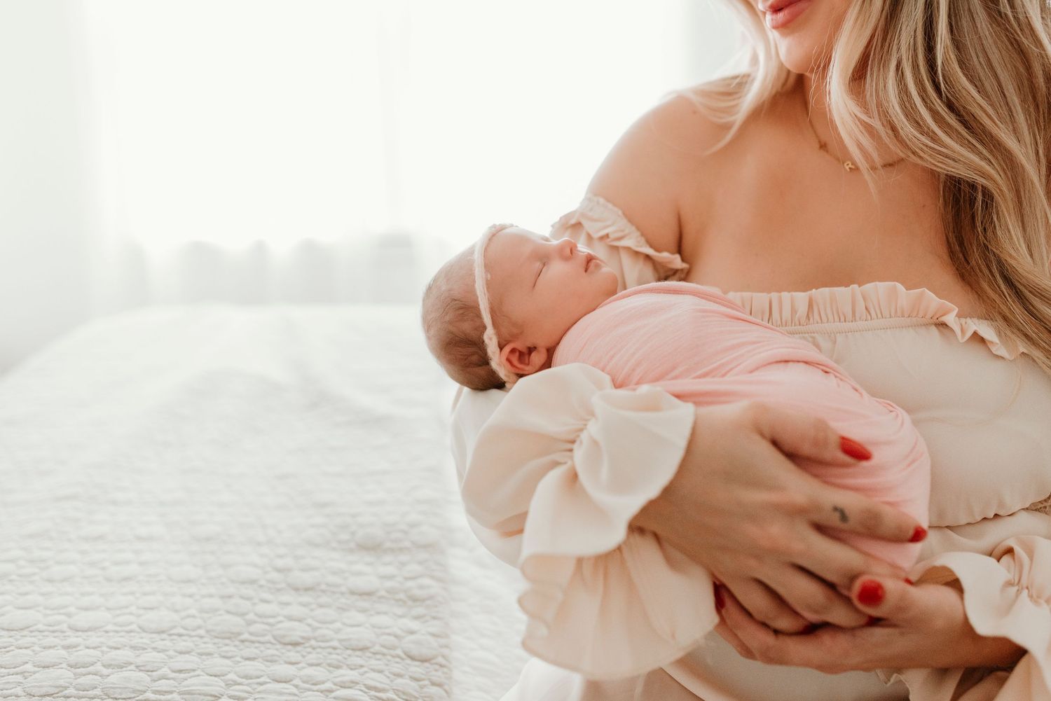 Mother in cream off-shoulder dress tenderly holds sleeping newborn baby dressed in pink.