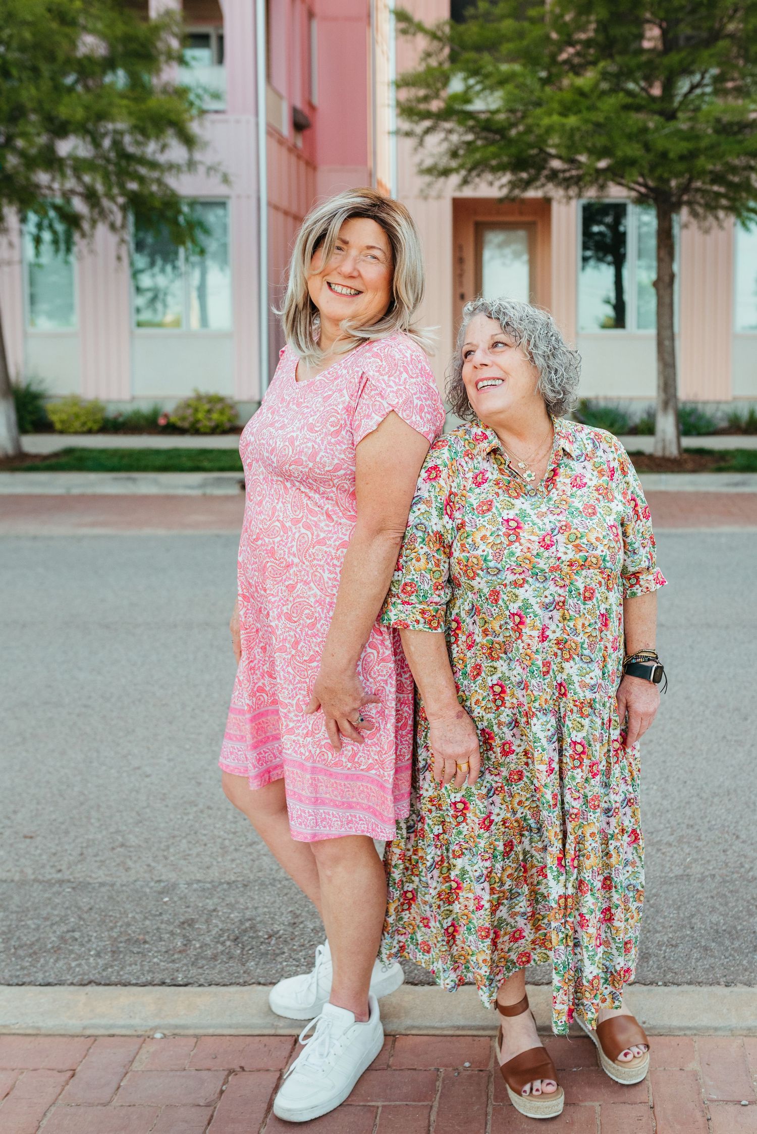 Two smiling women in pink and floral dresses standing together on a sidewalk in an urban setting.