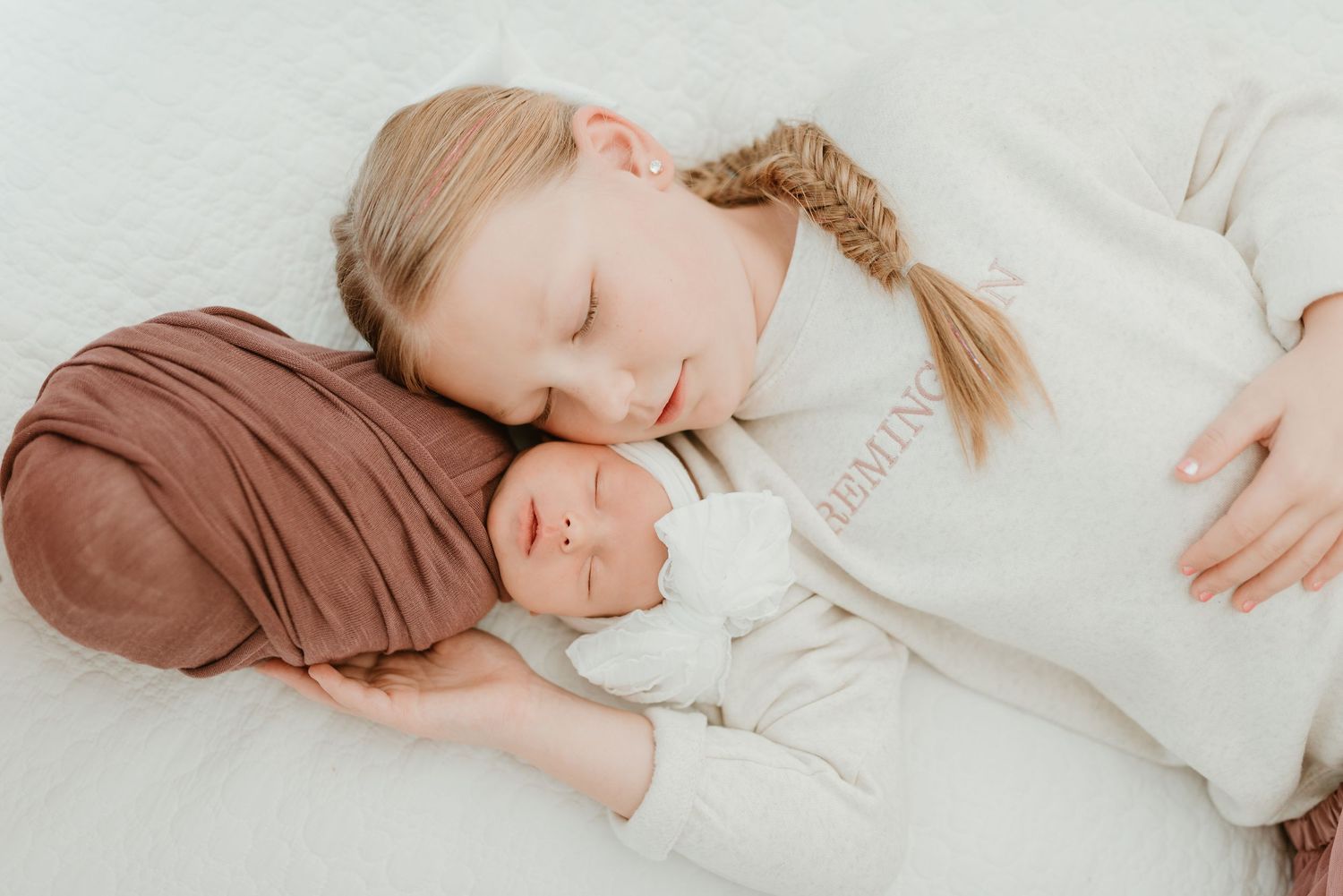 Young girl rests her head beside sleeping newborn sibling on white blanket in soft light.