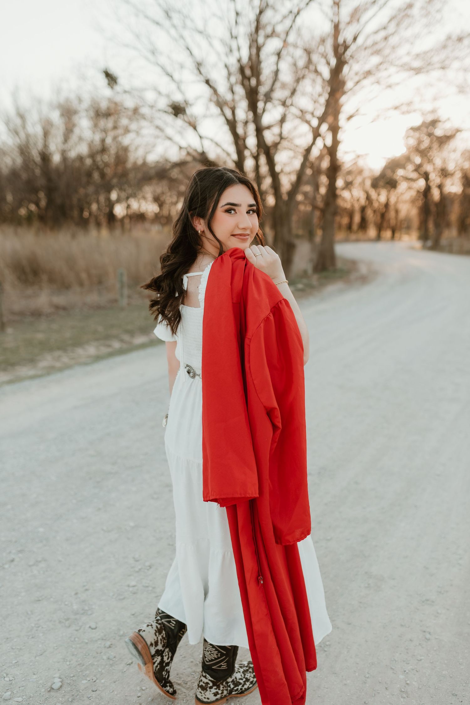Smiling young woman in white dress holding red blazer on a rural dirt road at sunset.