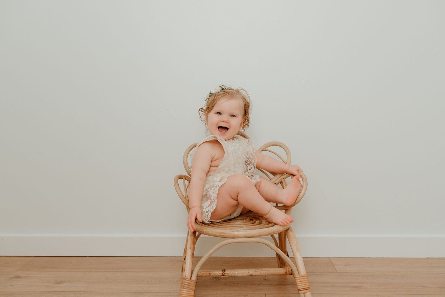 Smiling toddler girl in cream romper sitting in rattan chair against neutral white background.