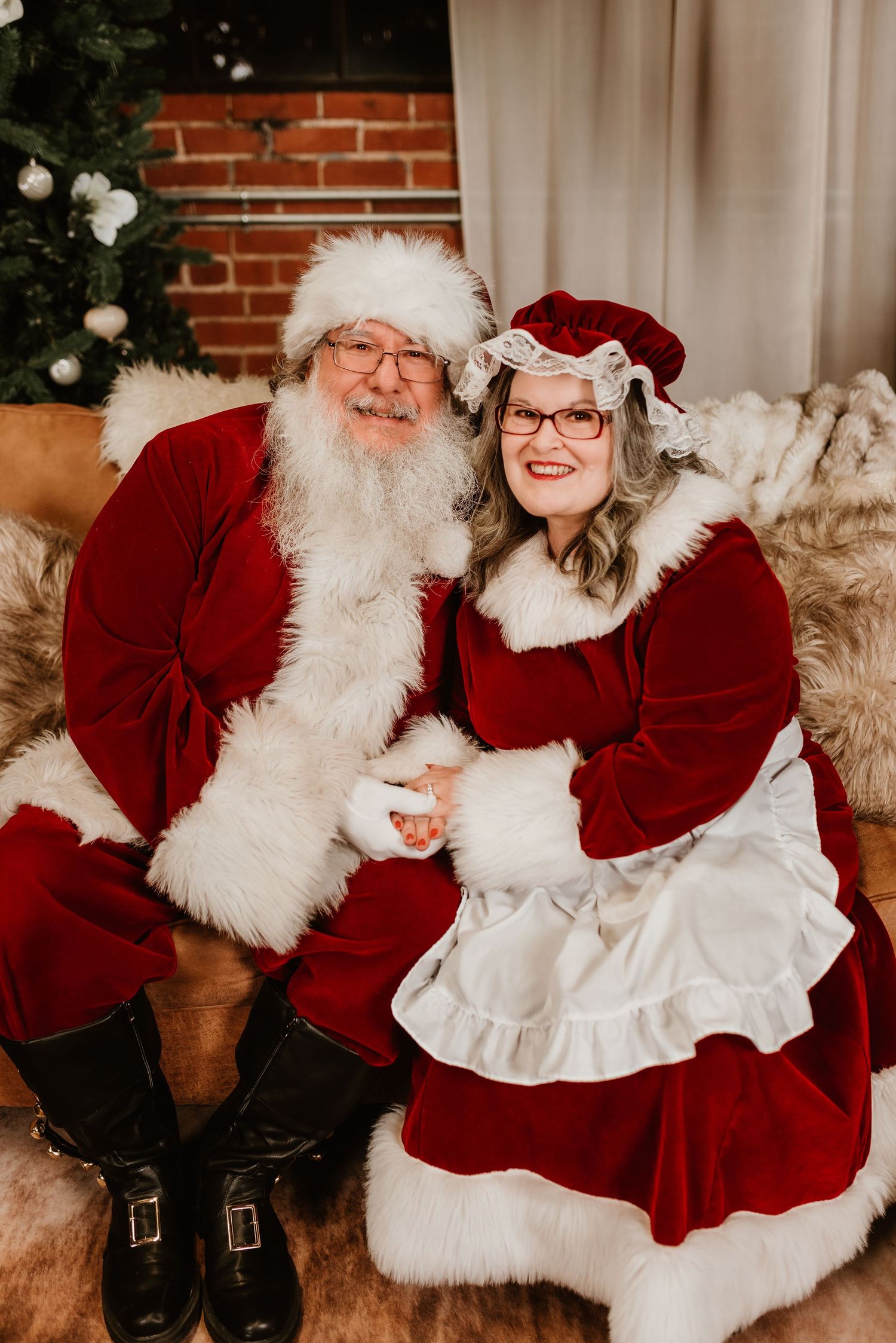 Santa Claus and Mrs. Claus sit together smiling in festive red velvet suits near a Christmas tree.