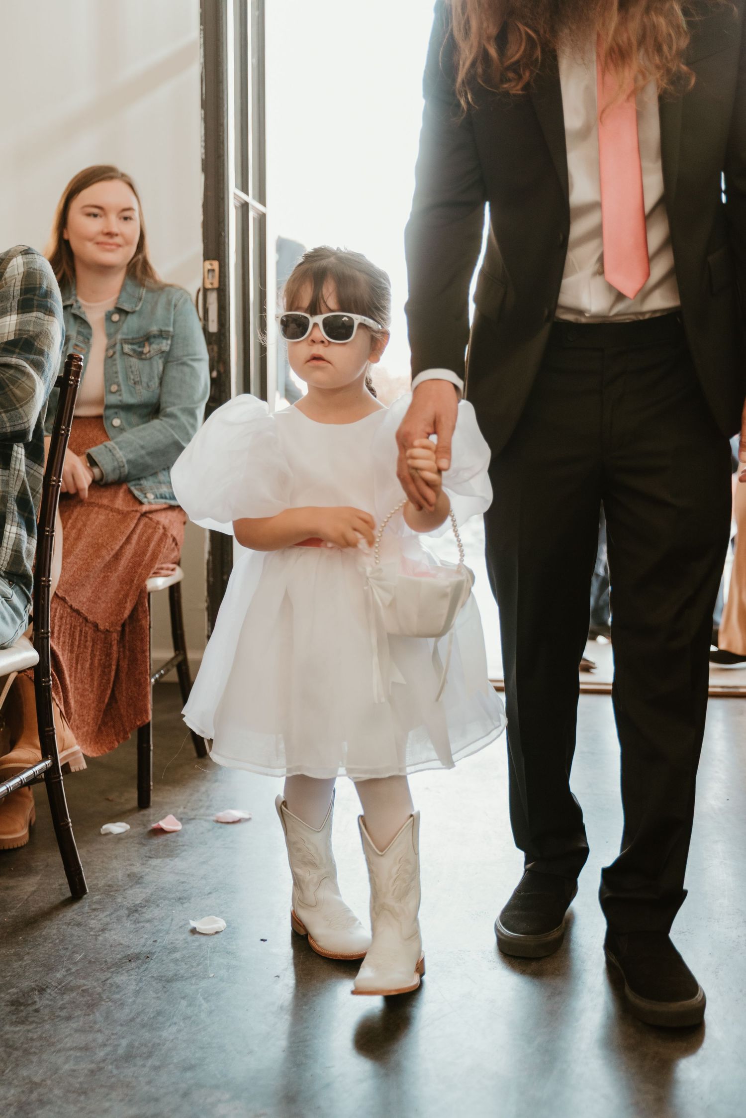 Adorable flower girl in white dress, sunglasses, and cowboy boots holding a basket at a wedding ceremony.