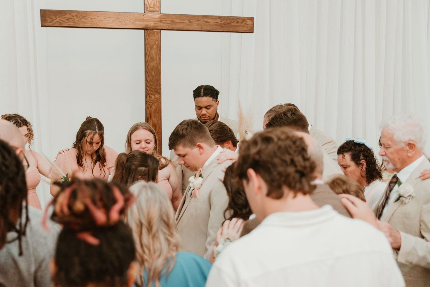Wedding party gathers in prayer around bride and groom before large wooden cross in white chapel.