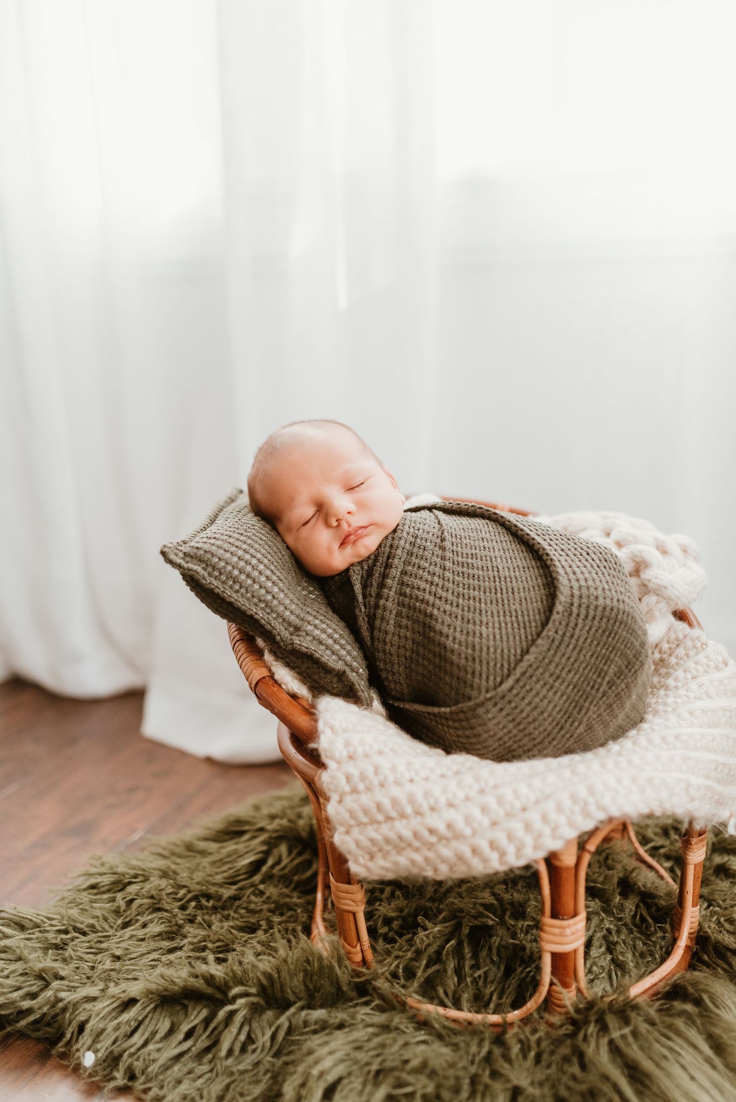 Peaceful newborn baby swaddled in olive wrap sleeping in small wicker rocking chair.