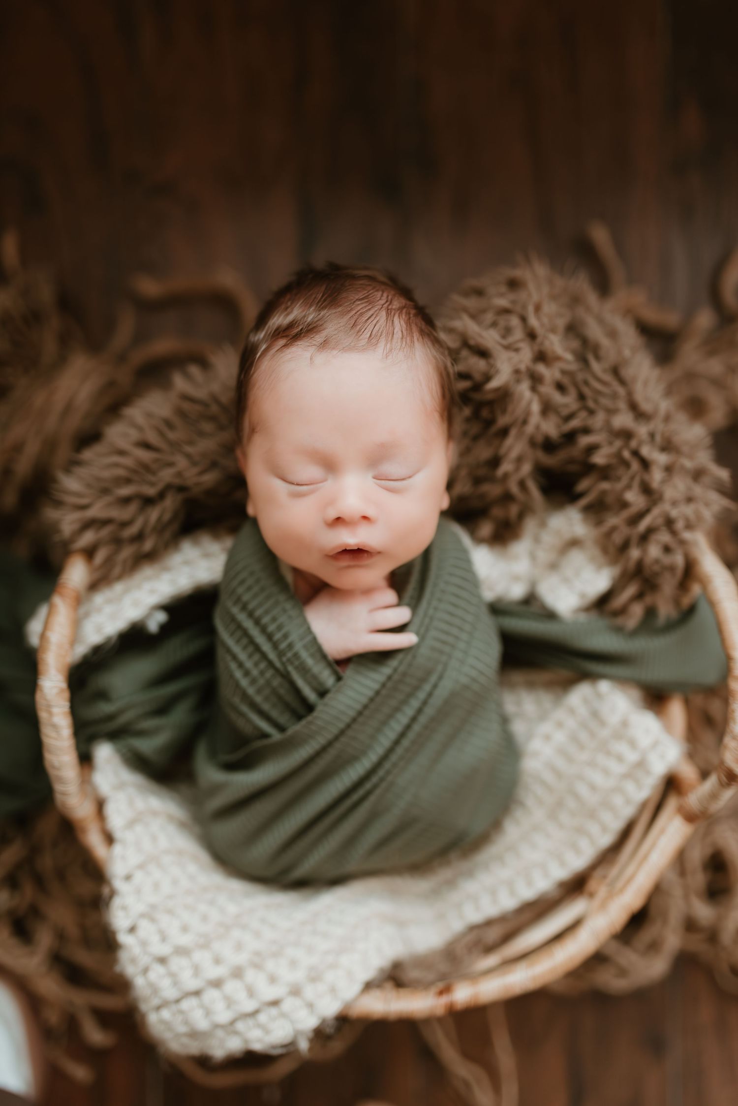 Sleeping newborn baby swaddled in green wrap nestled in a wicker basket with soft textures.