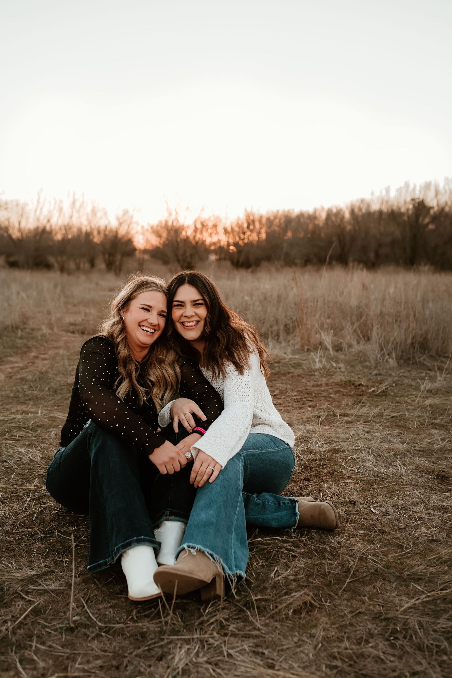 Two smiling women sit together laughing in a dry grass field during a warm sunset photo session.