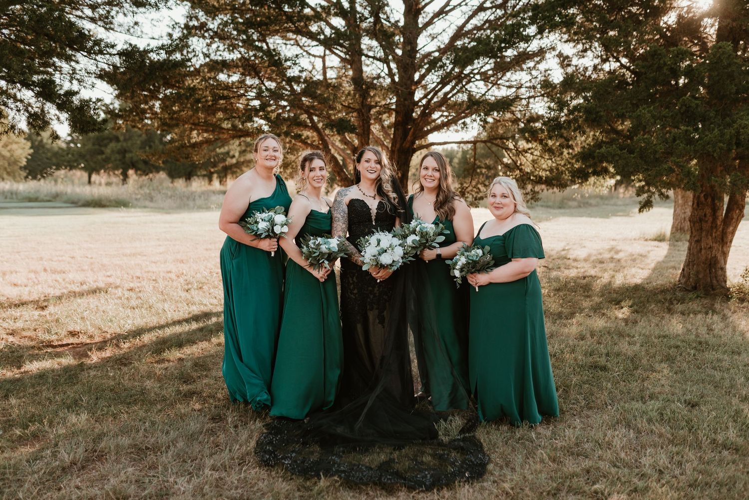 Bride in black gown surrounded by four bridesmaids in emerald green dresses holding white bouquets.