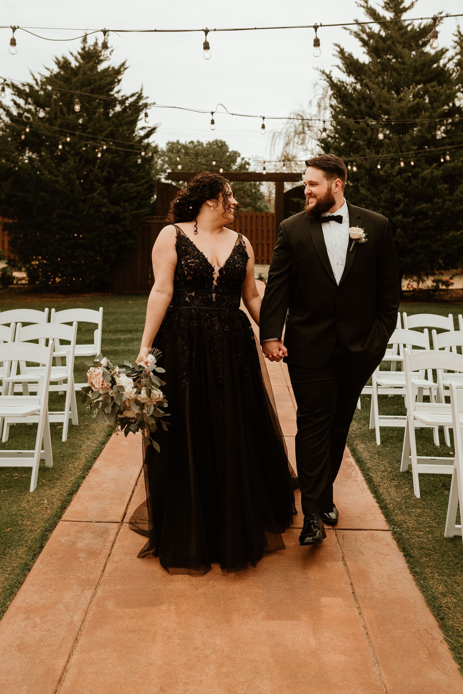 Bride in black wedding gown and groom in tuxedo walk hand in hand down outdoor aisle.