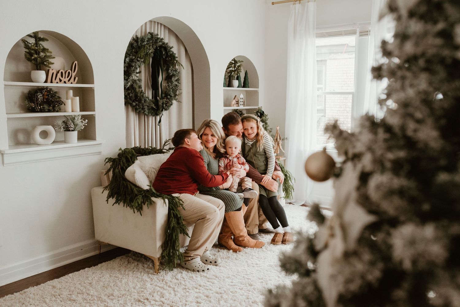 A family of five cuddles together on a white couch surrounded by Christmas wreaths and holiday decor.