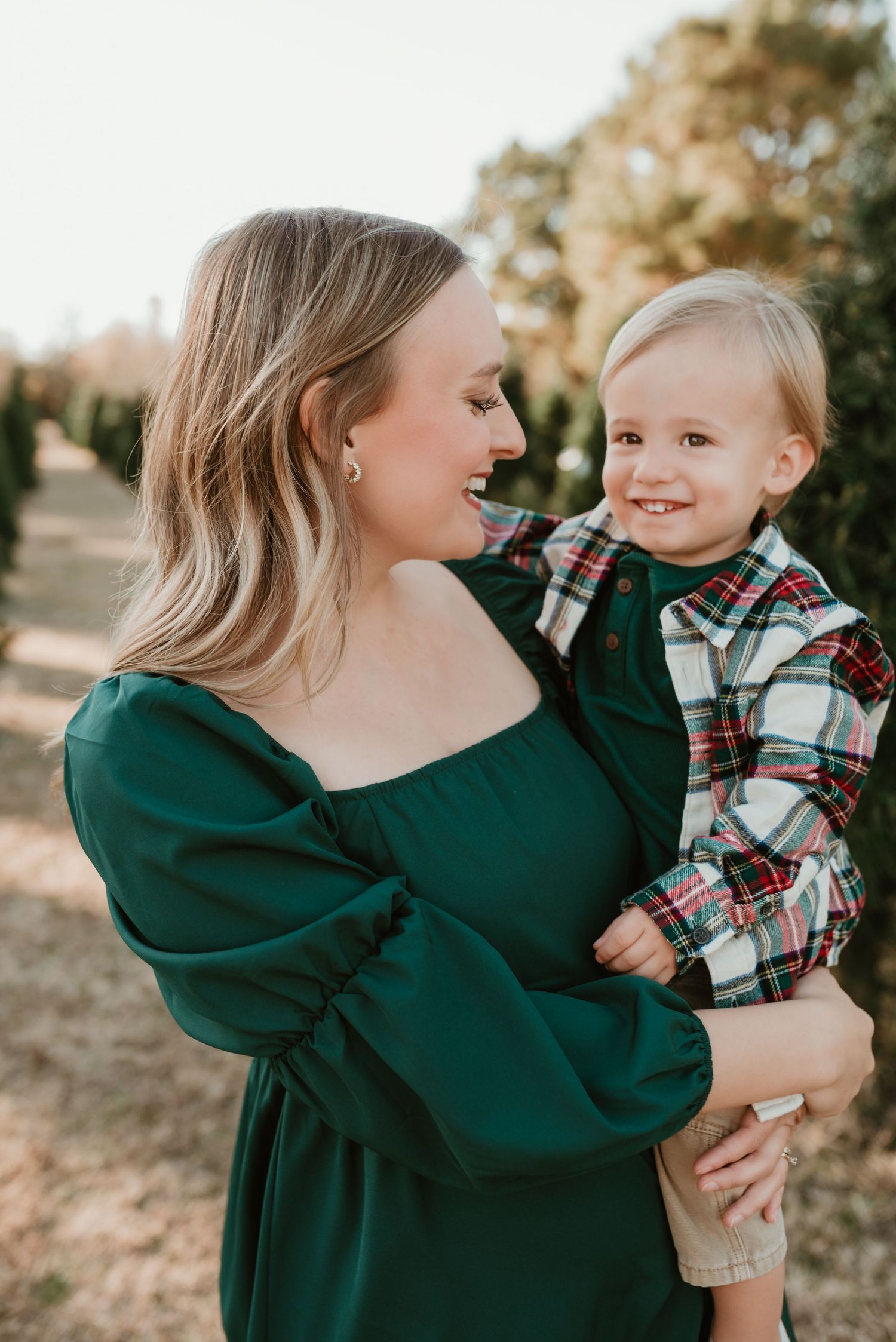 A smiling woman in a green dress holds a happy toddler boy in plaid shirt outdoors.