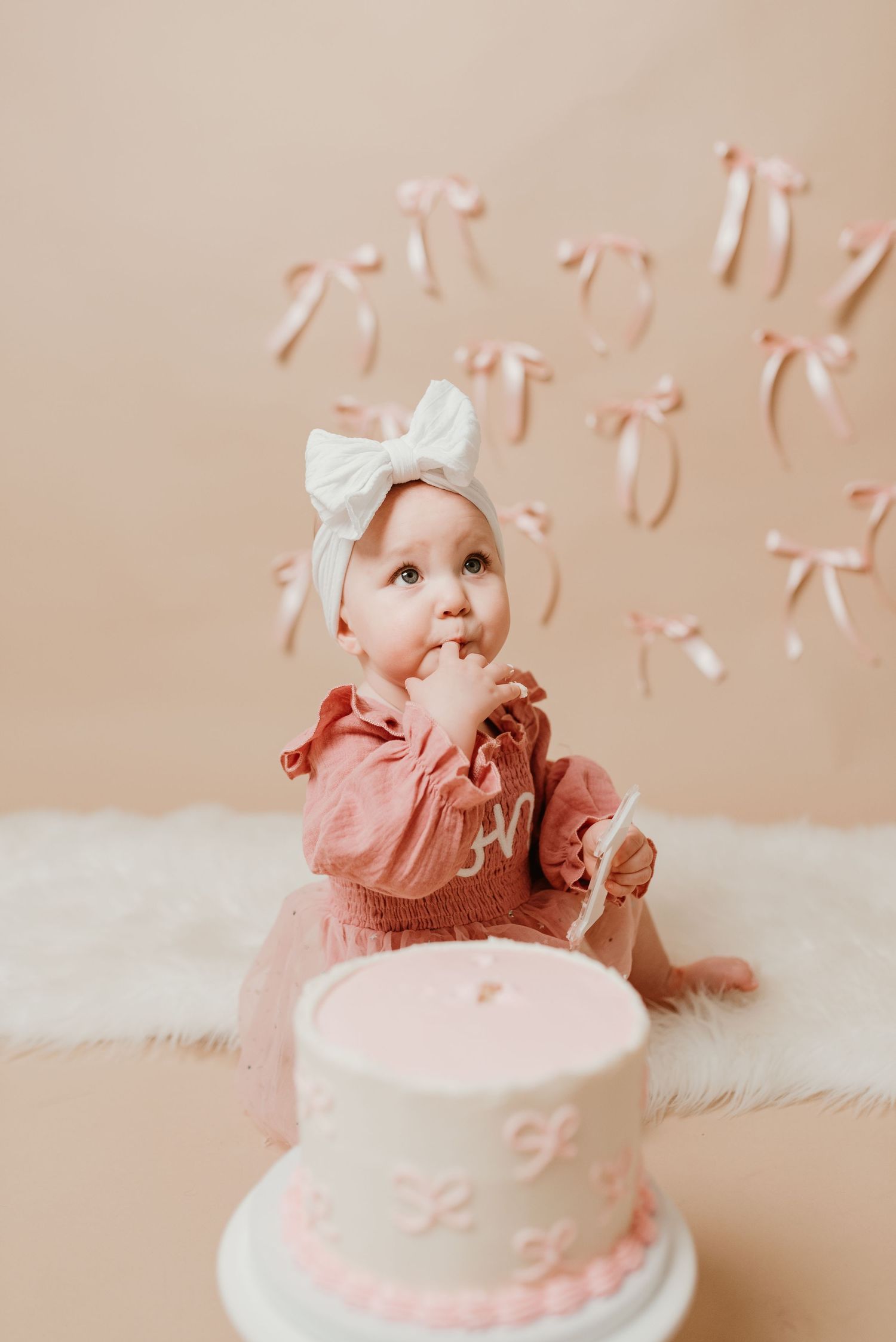 Baby girl in pink ruffle dress with white bow headband at cake smash photoshoot with decorated white cake.