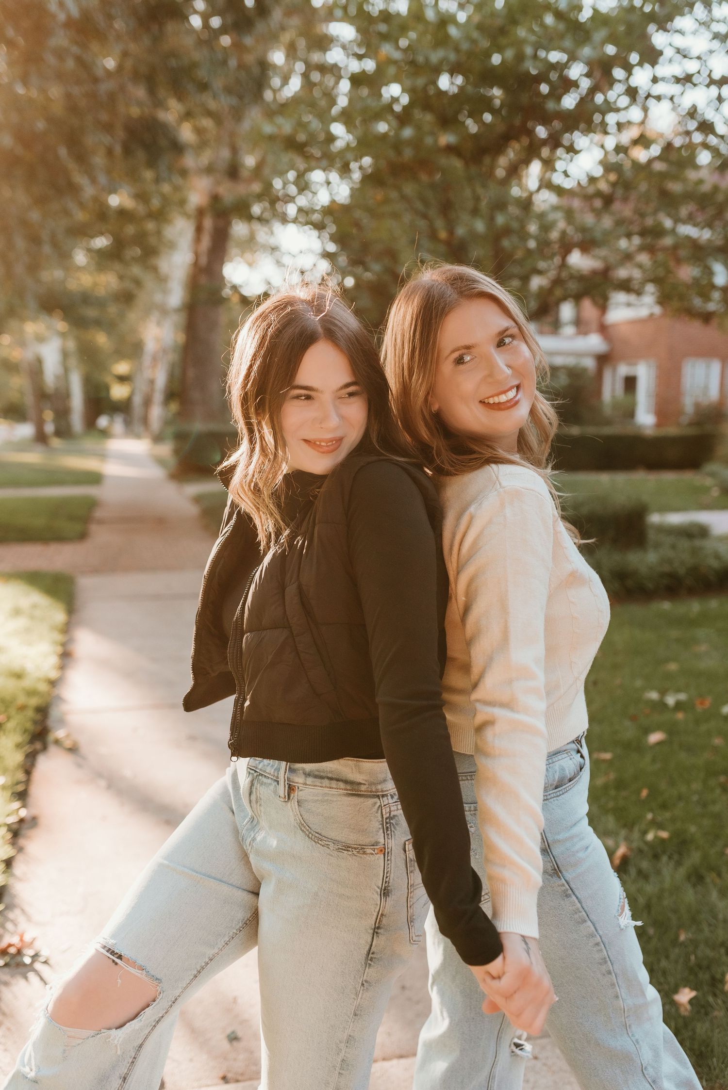 Two young women posing back-to-back in jeans and long-sleeve tops in a sunny park setting.