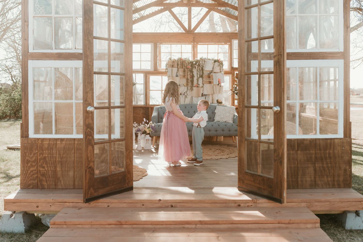 Couple holding hands walking through open French doors into a sunlit wooden greenhouse venue.