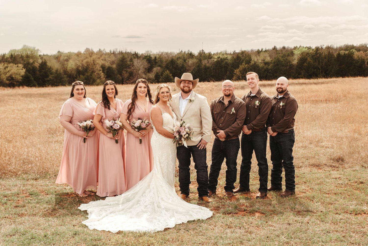 Wedding party poses in a golden field with bridesmaids in pink gowns and groomsmen in brown suits.
