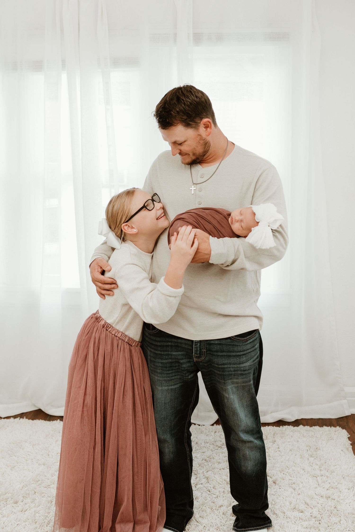 A father in a beige sweater and jeans holds a newborn while his young daughter in a pink tulle skirt embraces him warmly.