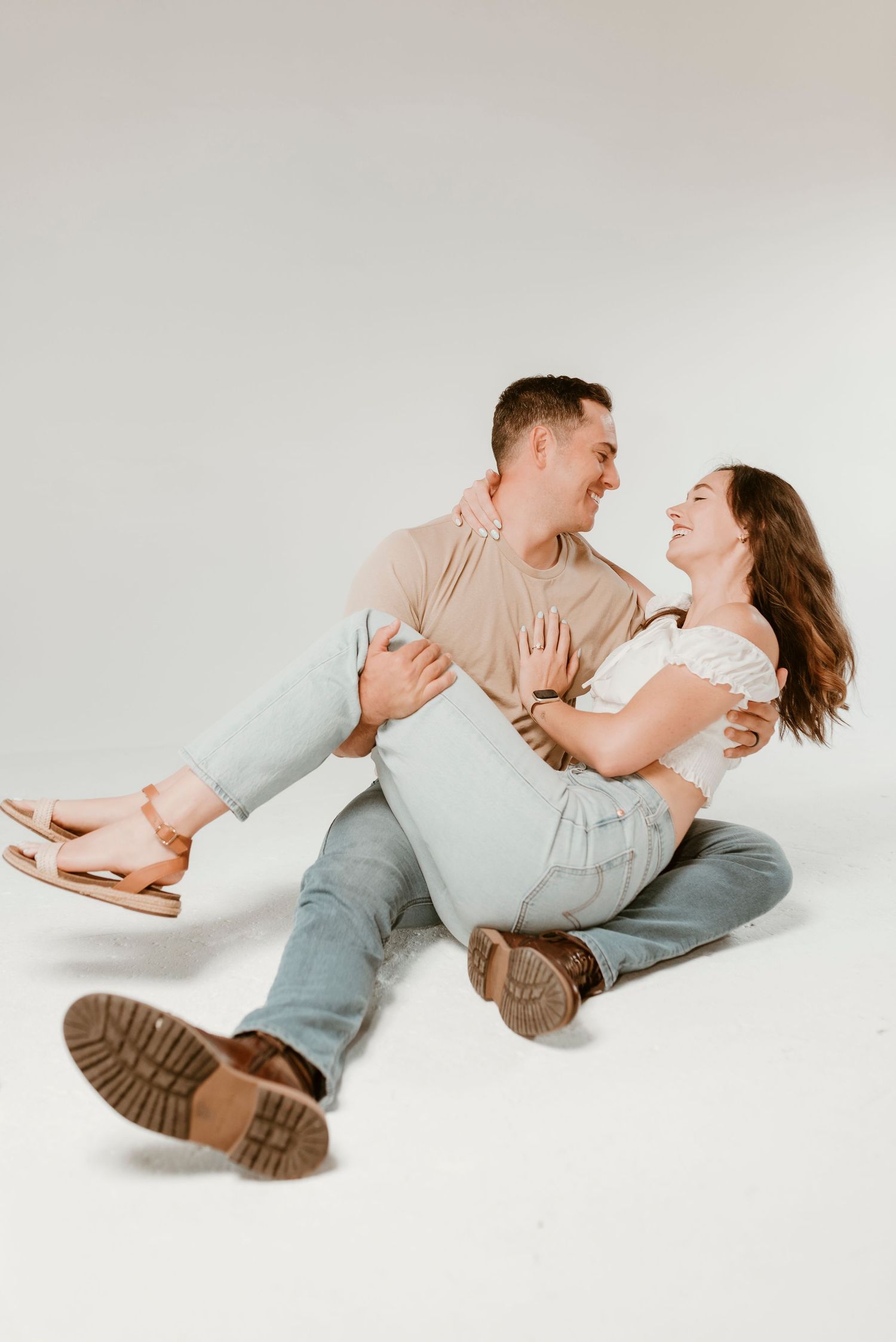Laughing couple sitting on a white studio floor, the man holding the woman in his lap.