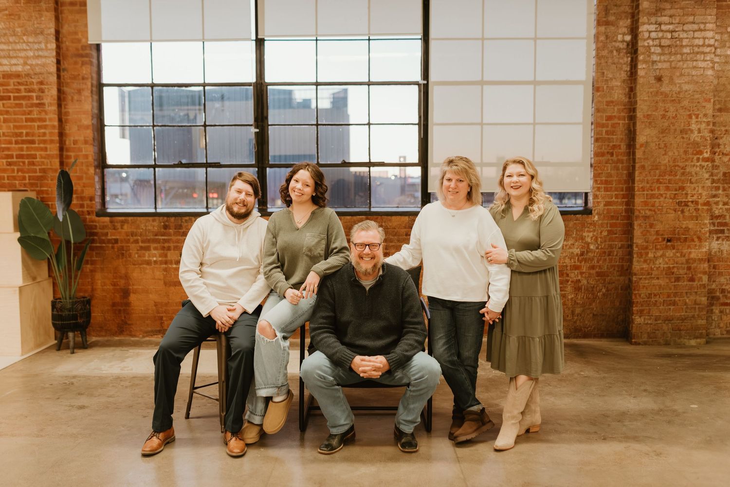 Five colleagues pose together in a modern brick-walled loft studio with large industrial windows.