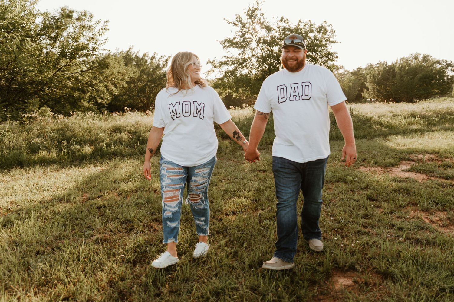 Couple wearing matching MOM and DAD shirts holding hands in a sunny green field.