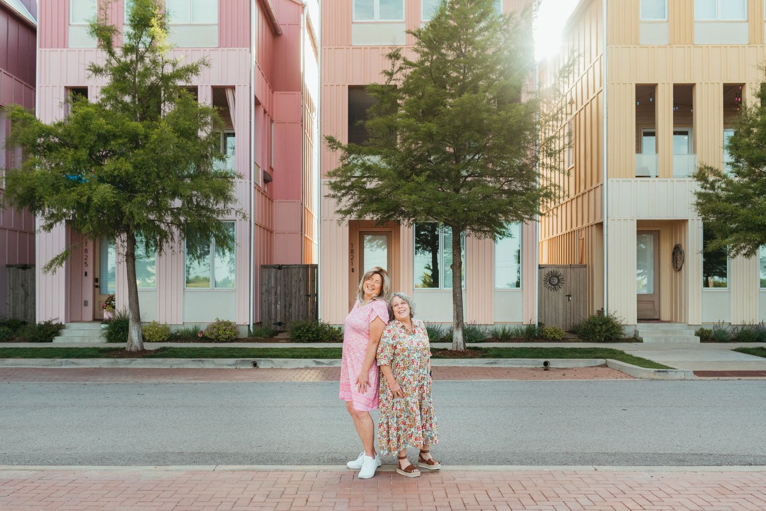 Two women in pink and floral dresses embracing on a brick street with colorful modern townhomes behind them.