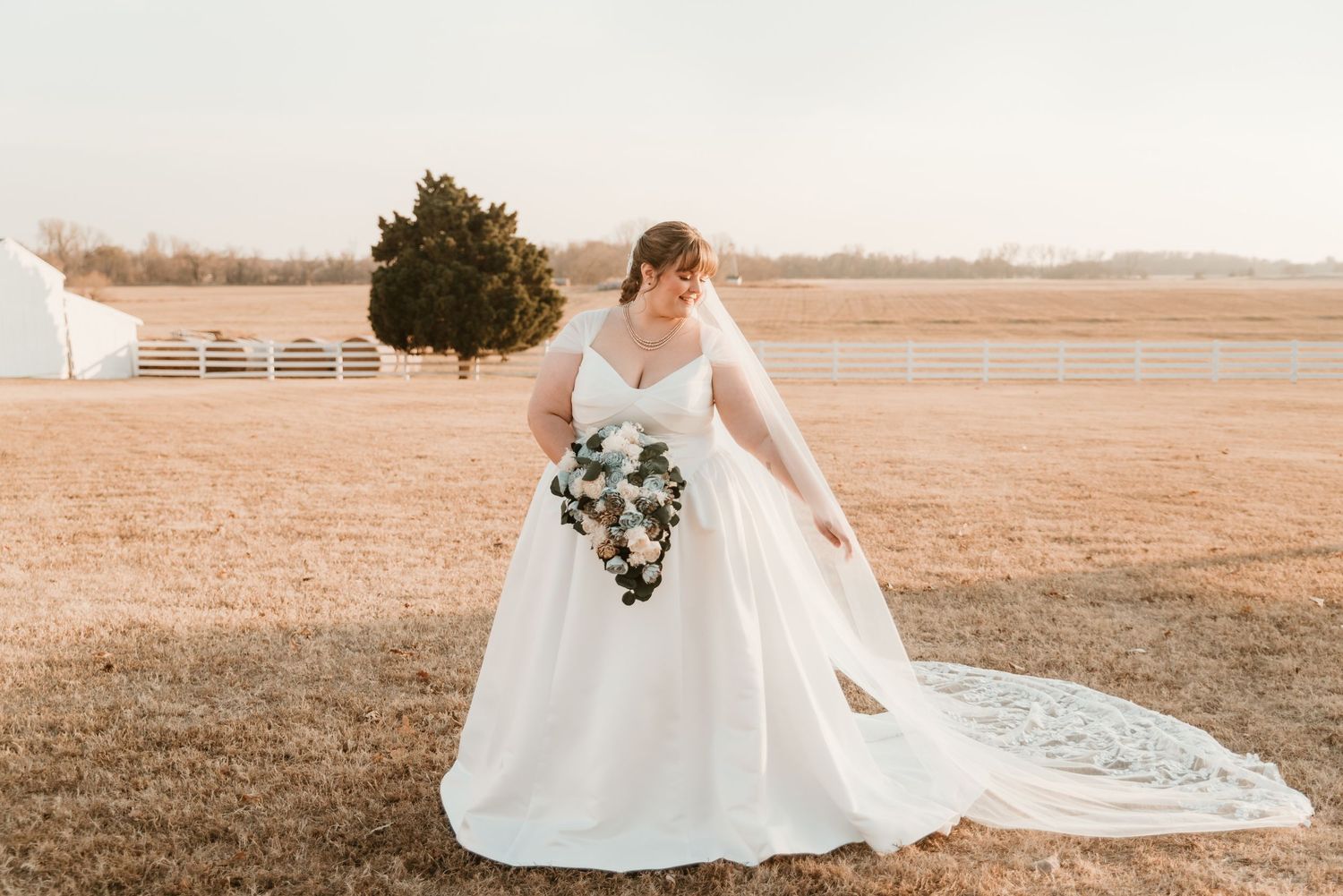 Bride in a classic A-line wedding gown with long train holding a bouquet on a rustic farm at sunset.