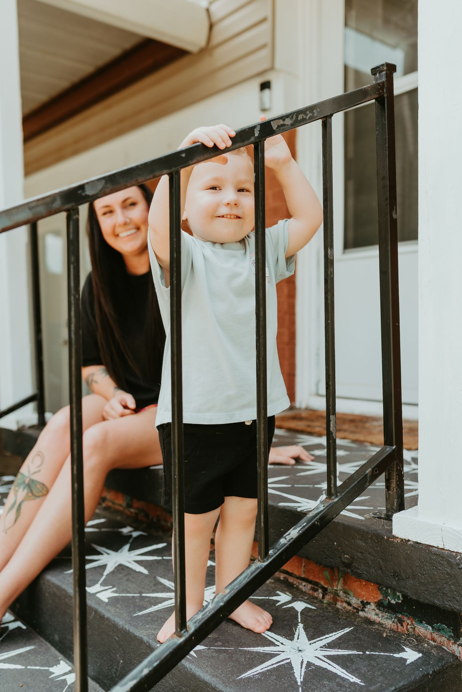 Happy toddler boy hangs on black iron porch railing while tattooed mother smiles from decorated front steps.