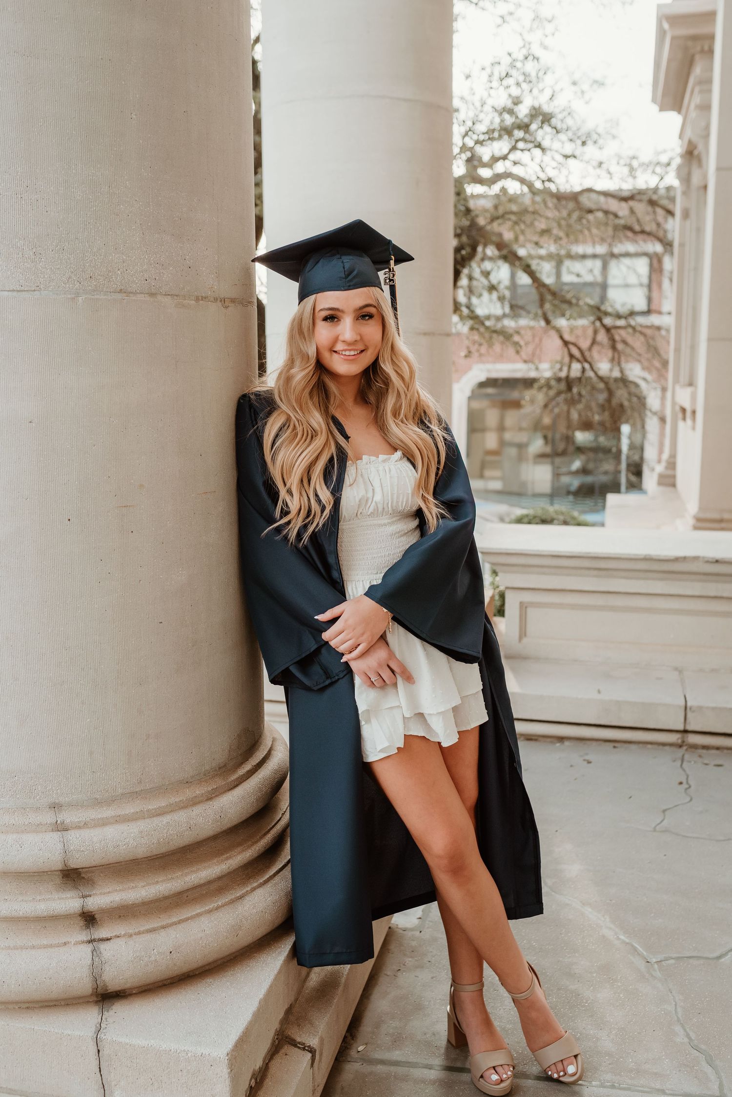 A blonde graduate in cap and gown leans against a column in a white dress and heels.