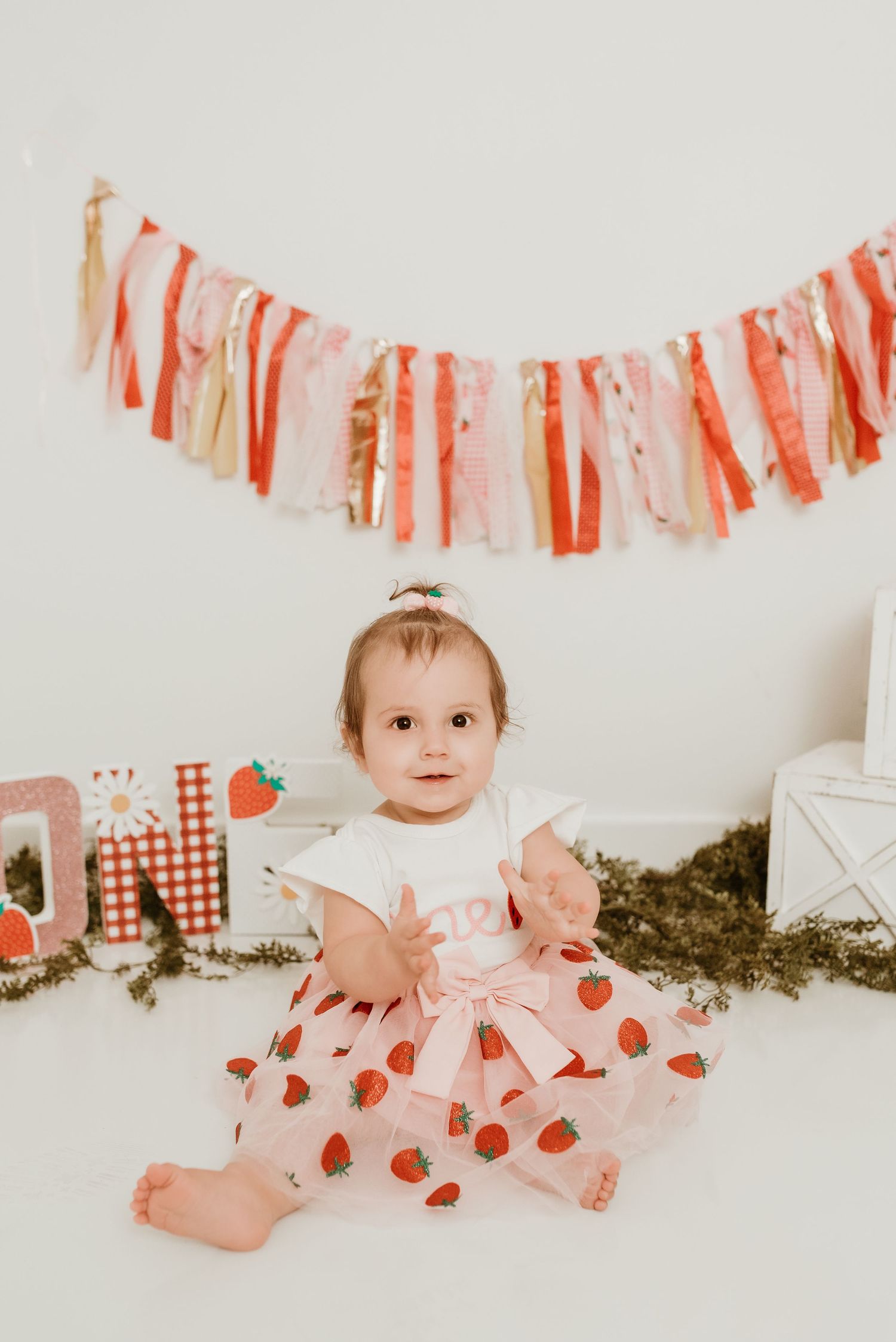 Baby girl in strawberry dress sitting at first birthday cake smash setup with colorful fabric banner.