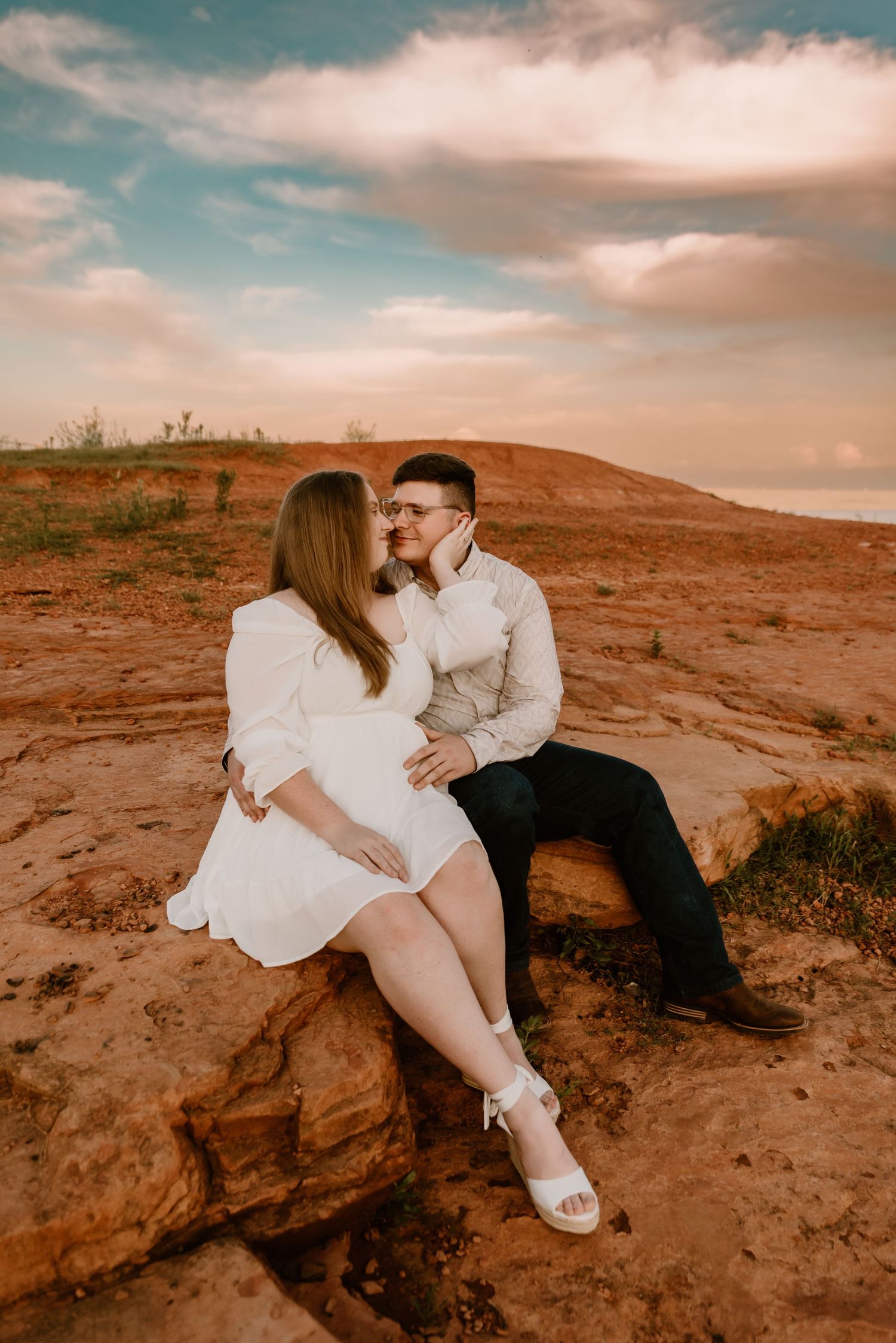A couple shares a romantic kiss while sitting on red rock formations under a dramatic cloudy sky.