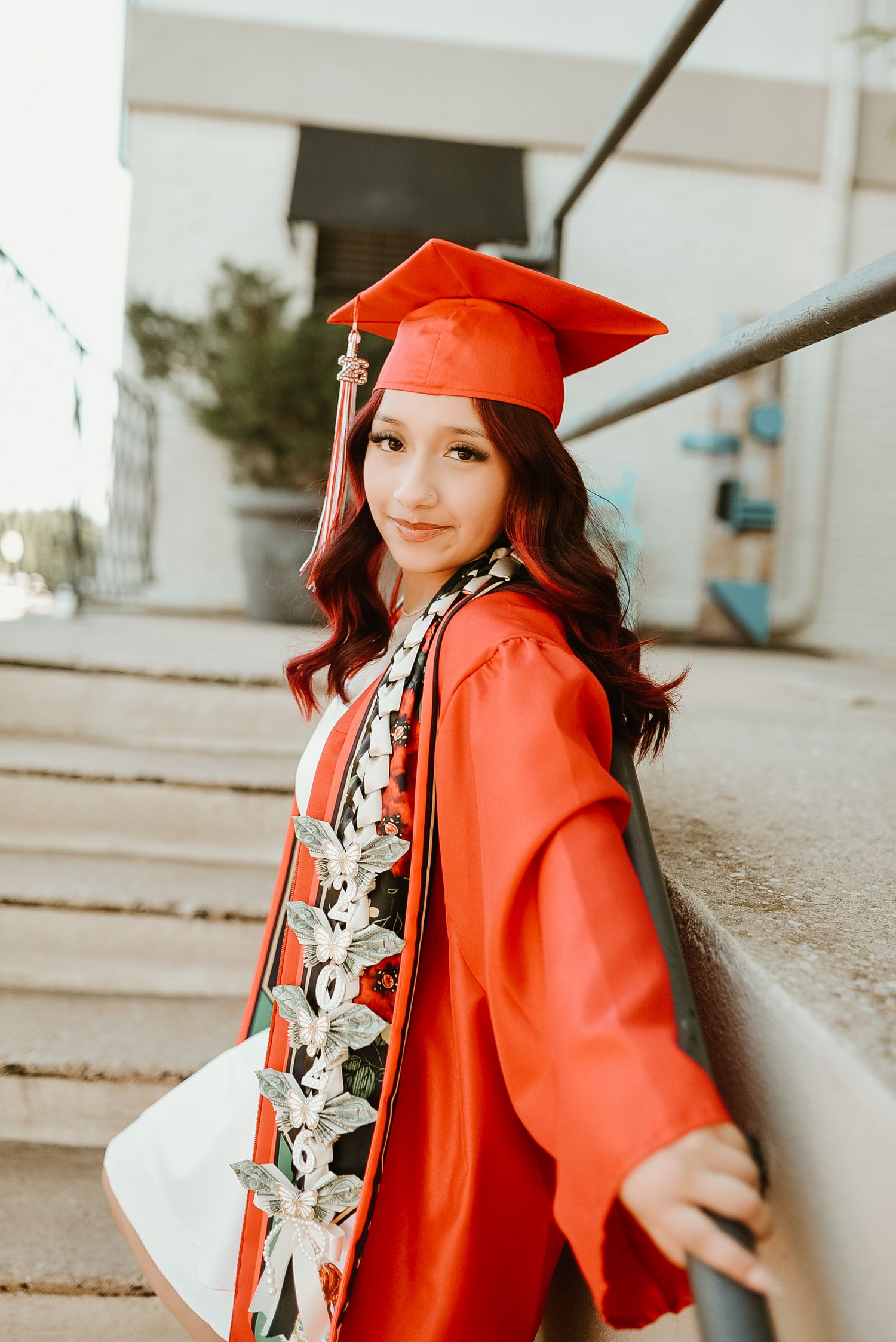 Young woman in red graduation cap and gown smiling on steps, wearing decorative sash, holding diploma.