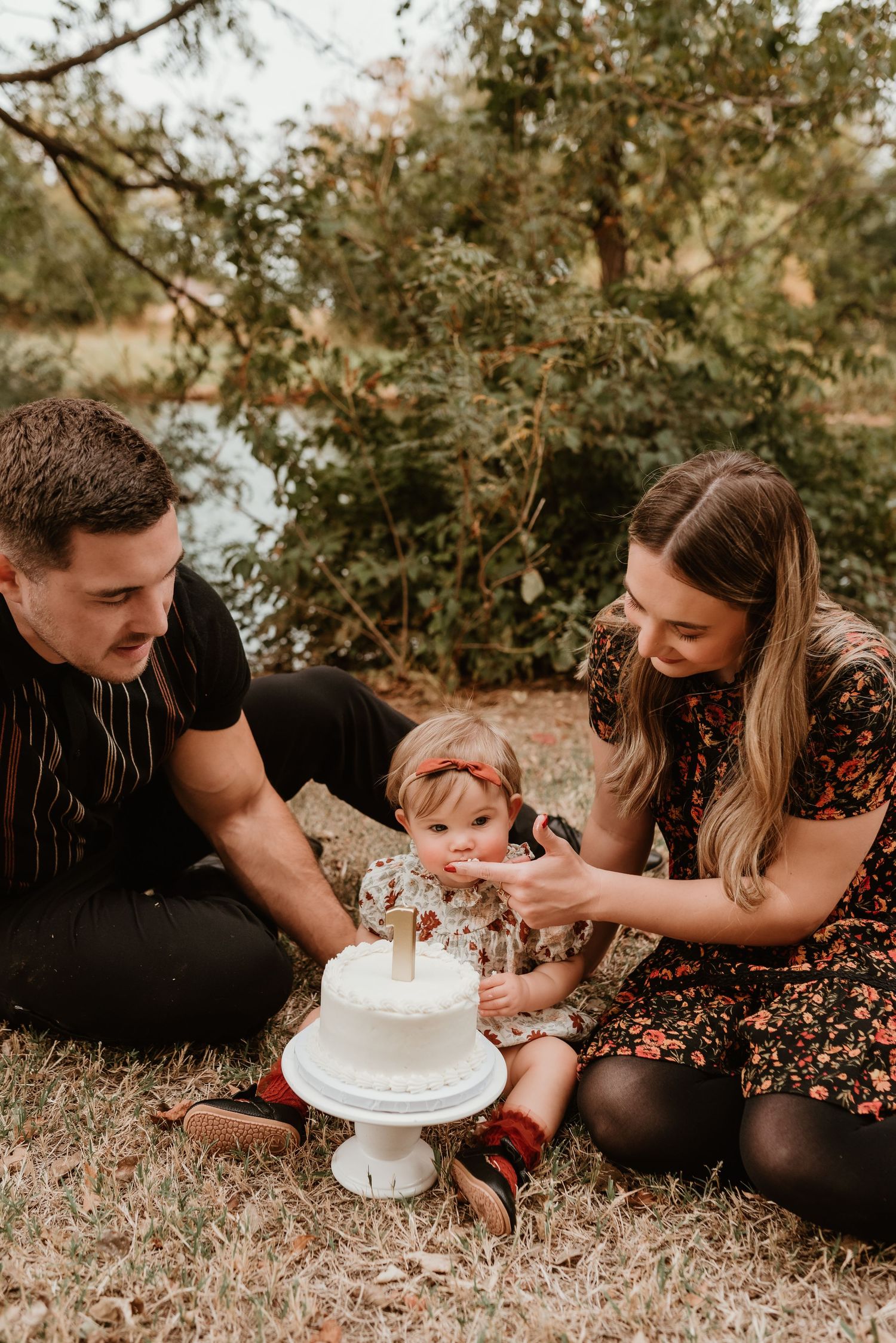 Parents sit with their baby girl on grass celebrating her first birthday with a white cake.
