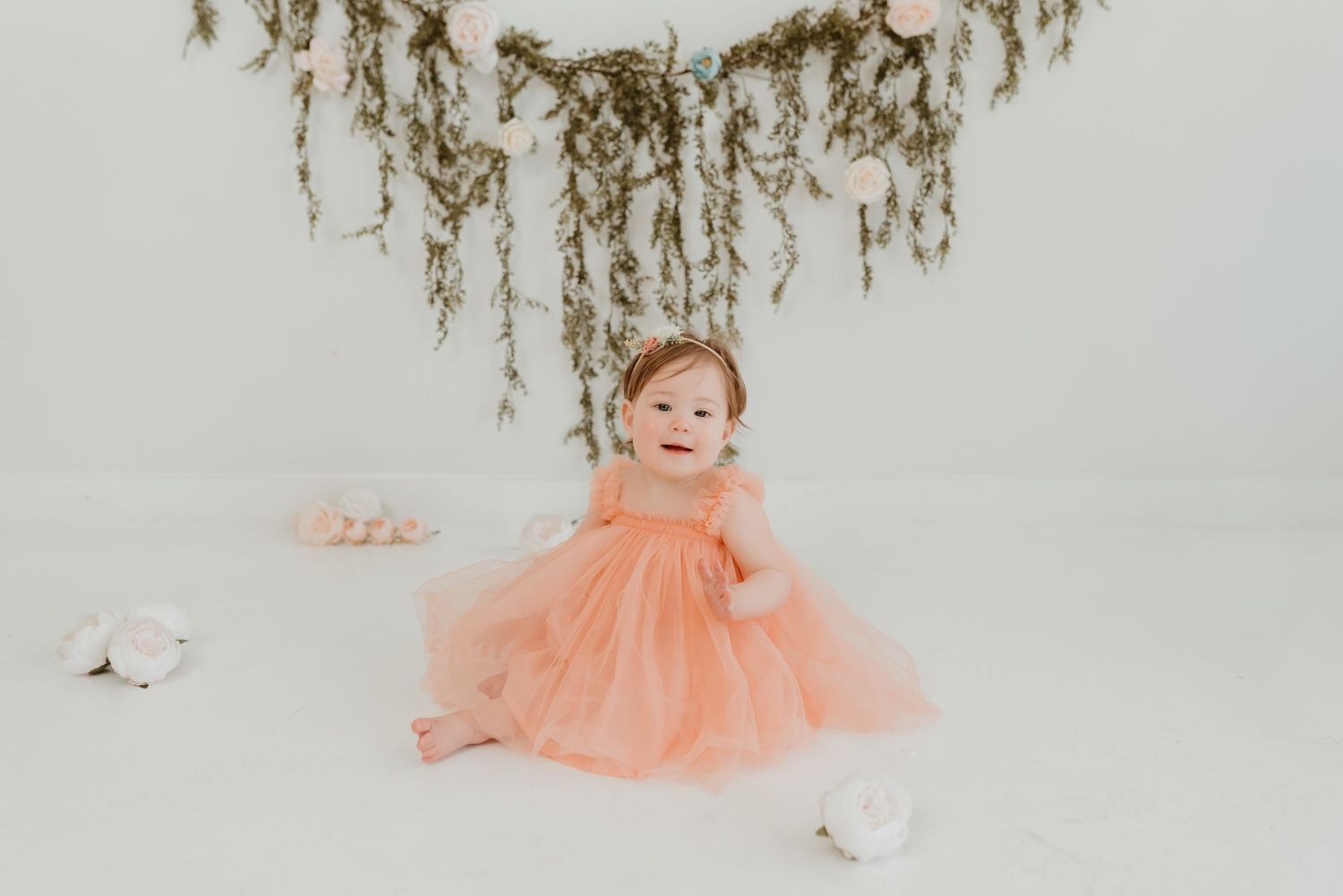 Smiling baby girl in peach tulle dress sitting on white floor under hanging greenery floral backdrop.