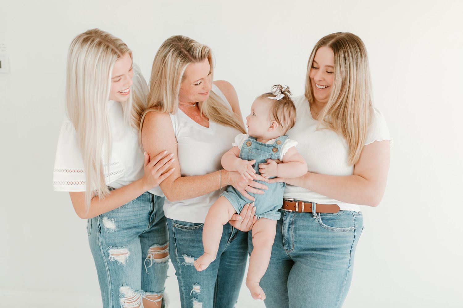 Three blonde women in white tops and jeans smile at an adorable baby in a bright studio photo.
