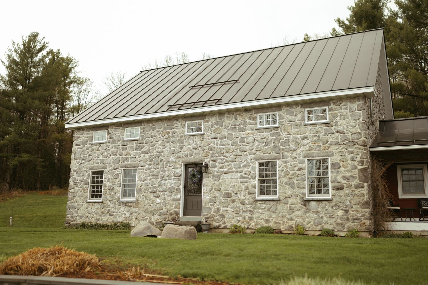 A historic stone barn with metal roof and multiple windows sits nestled among evergreen trees on rolling farmland.