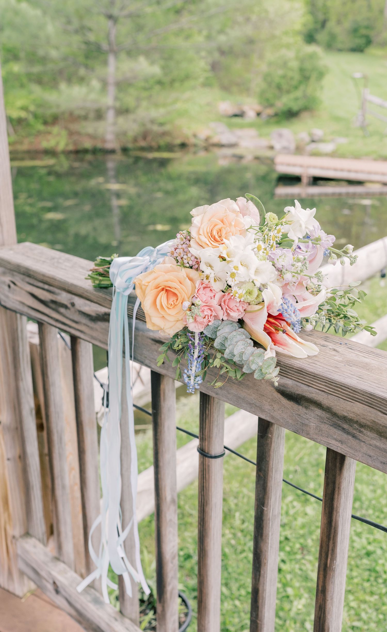 Delicate floral arrangements with peach roses and white flowers adorn a wooden railing overlooking a serene pond.