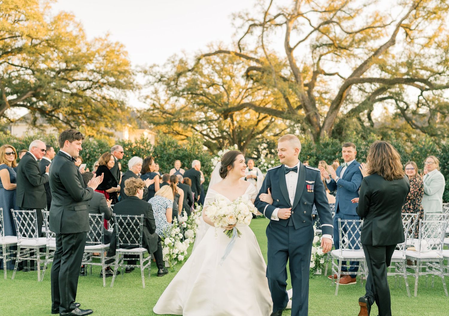 Wedding ceremony guests mingle outdoors during golden hour beneath autumn trees at an elegant garden venue.