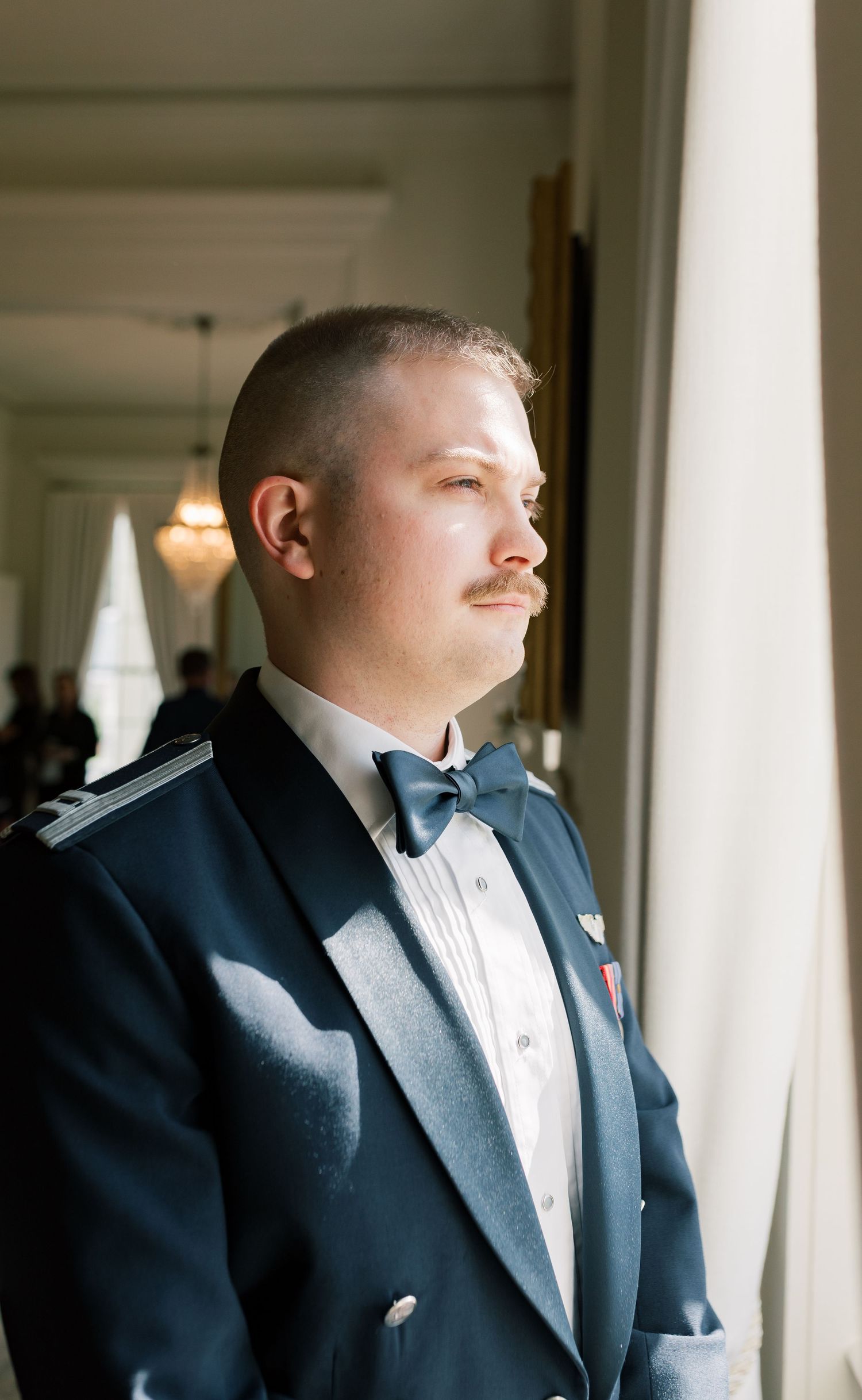 A groom in a black tuxedo and white bowtie stands by a window in natural light during a wedding.