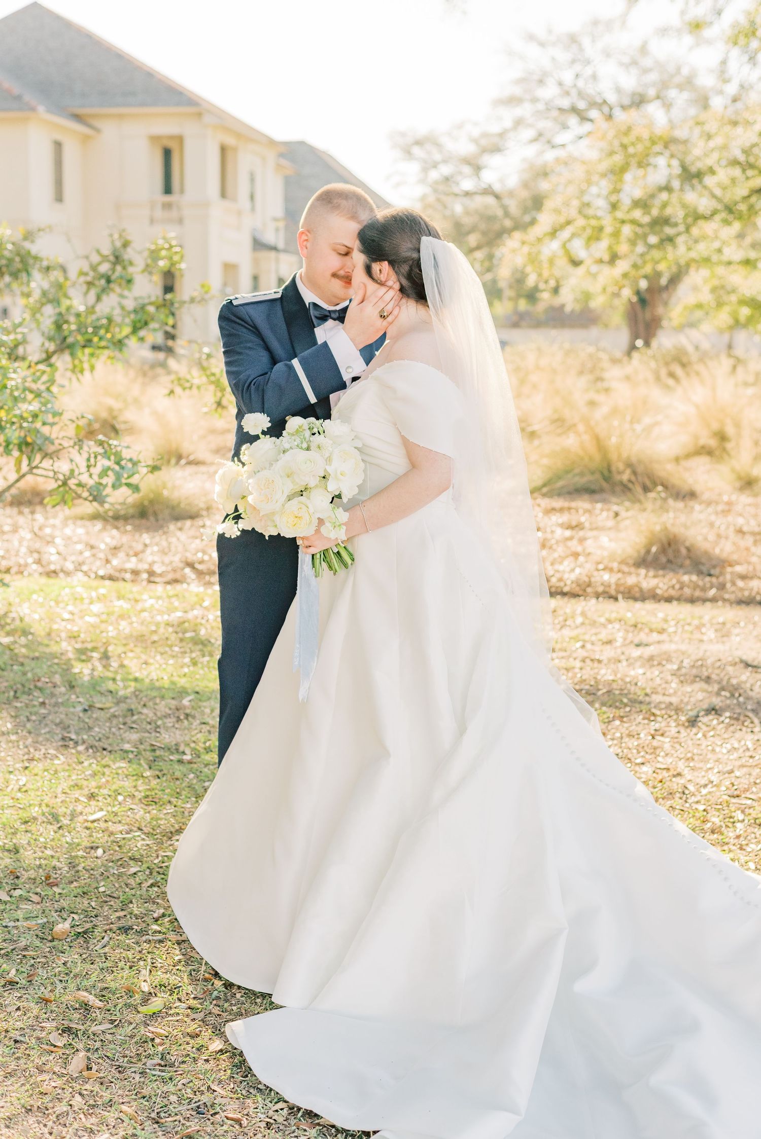 A romantic wedding couple embraces in a sun-drenched outdoor setting with elegant white dress and navy suit.