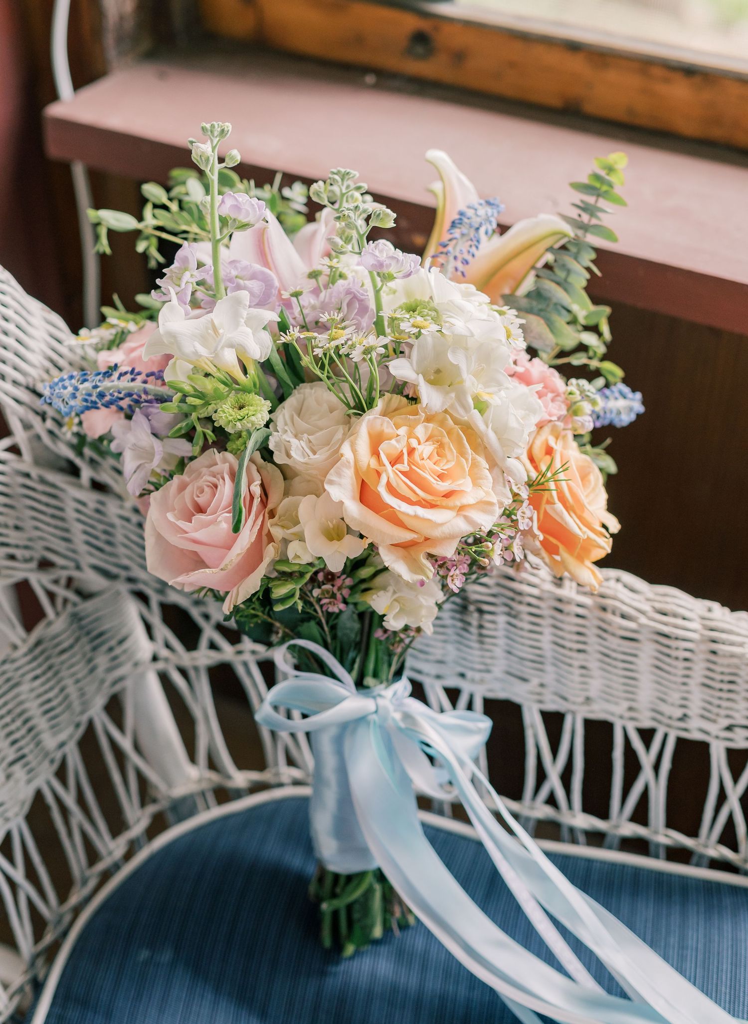 Wedding bouquets with peach and pink roses arranged with delicate ribbons on white wicker furniture.